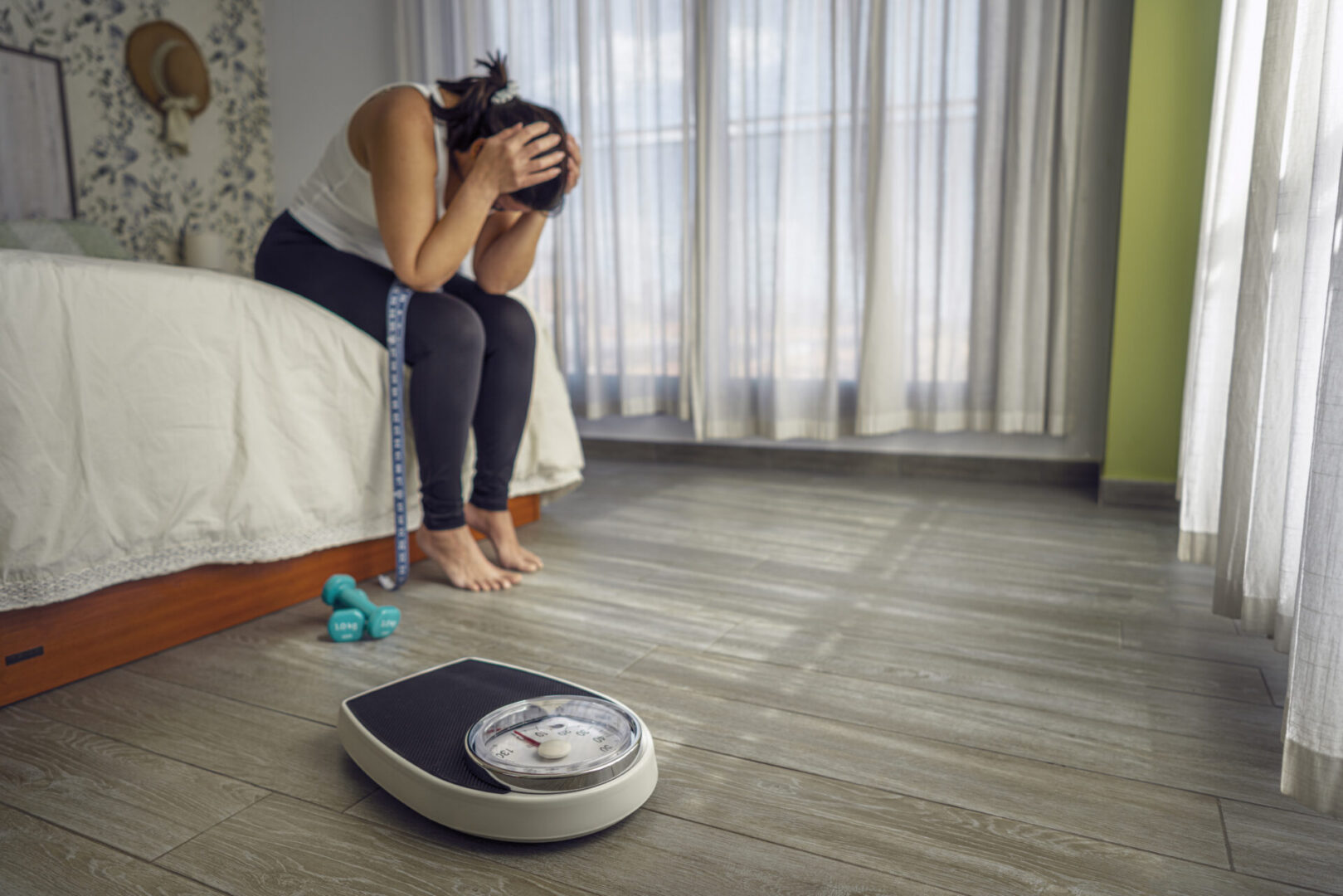 Overweight woman sitting on the bed head in hands worried about her obesity. Bathroom scale and dumbbells are on foreground. High resolution 42Mp indoors digital capture taken with SONY A7rII and Zeiss Batis 25mm F2.0 lens
