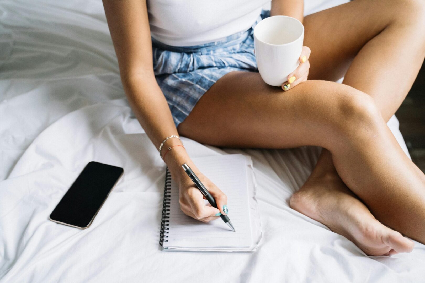 Person writing in notebook on bed with coffee