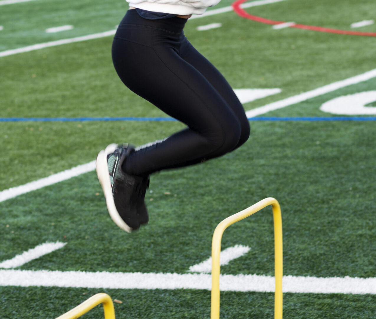 A high school girl is jumping over plastic yellow hurdles on a turf field waring black spandex and a white sweatshirt during track and field practice.