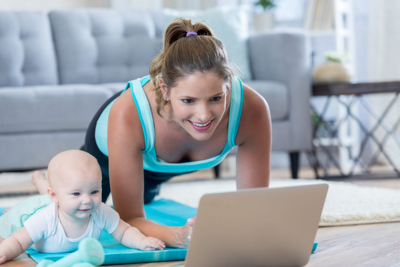 Cheerful Caucasian woman  works out while watching an exercise video on her laptop. Her infant daughter is lying beside her on a yoga mat. The woman is in a plank position.