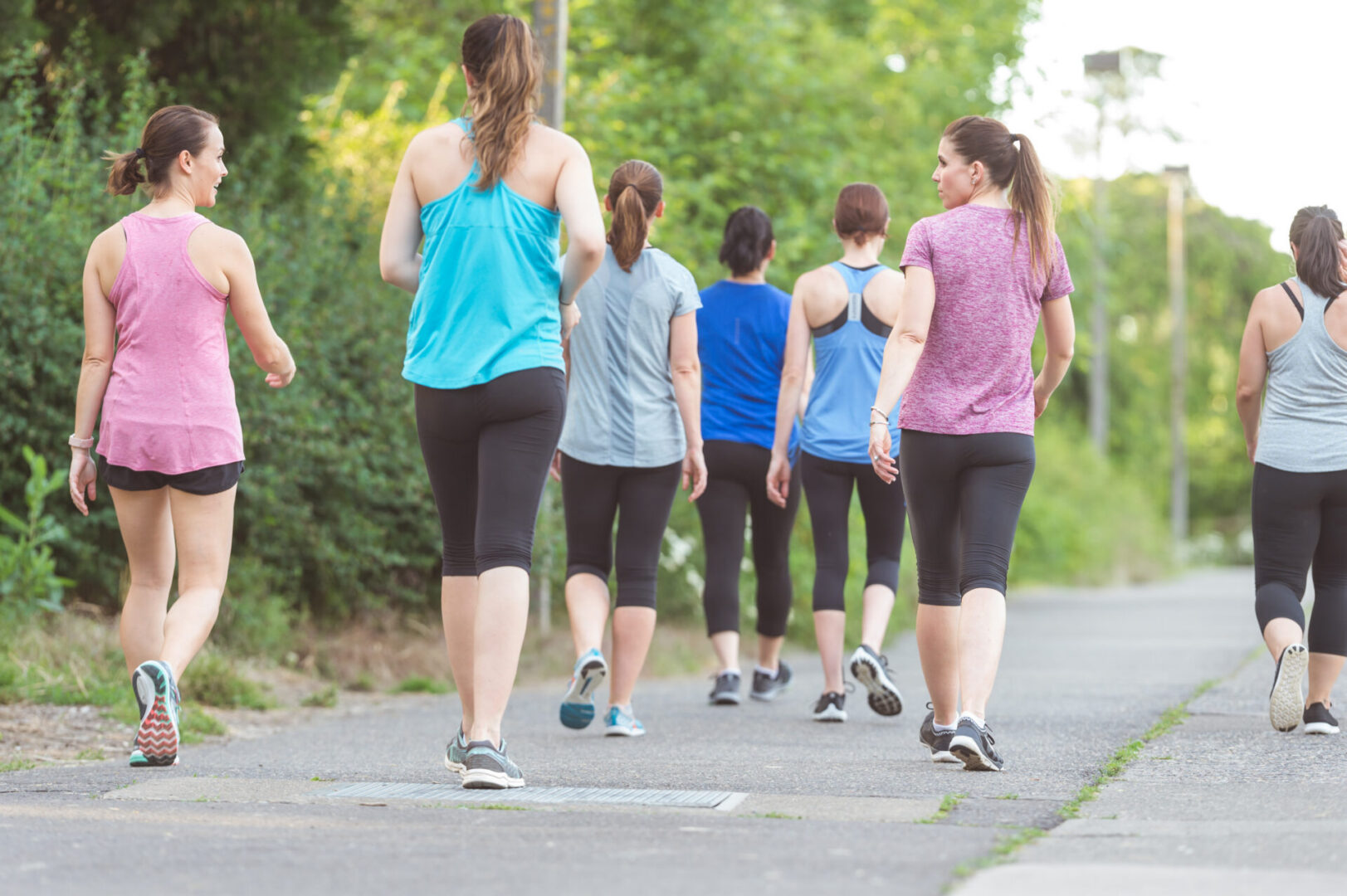 Group of women participate in a race run/walk to raise money for breast cancer research. They are wearing race bibs and are on a sidewalk, with trees on the left side. The shot is from behind them as they walk along the sidewalk.